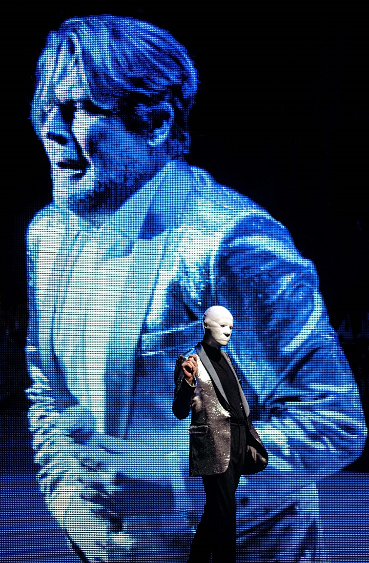 Mężczyzna na scenie w białej masce, drugi wyświetlany na projekcji, bez maski, w garniturze, odwróceni w dwie strony