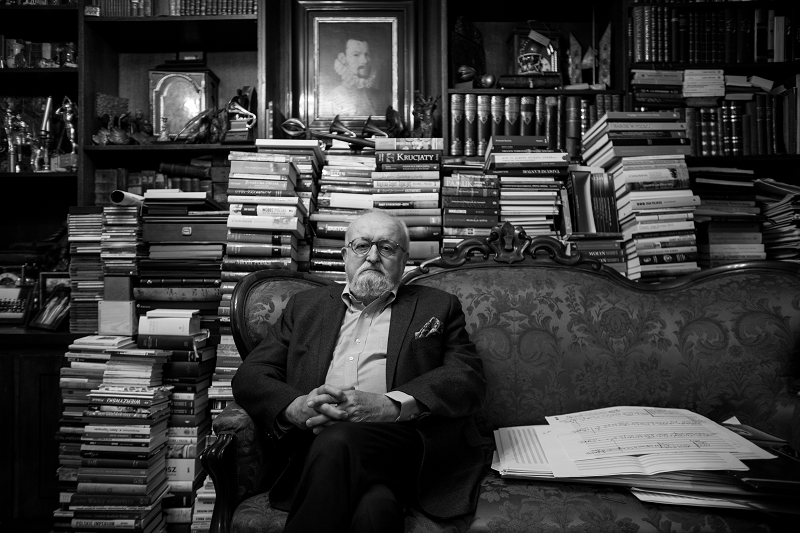 Krzysztof Penderecki sits on a settee surrounded by stacks of books