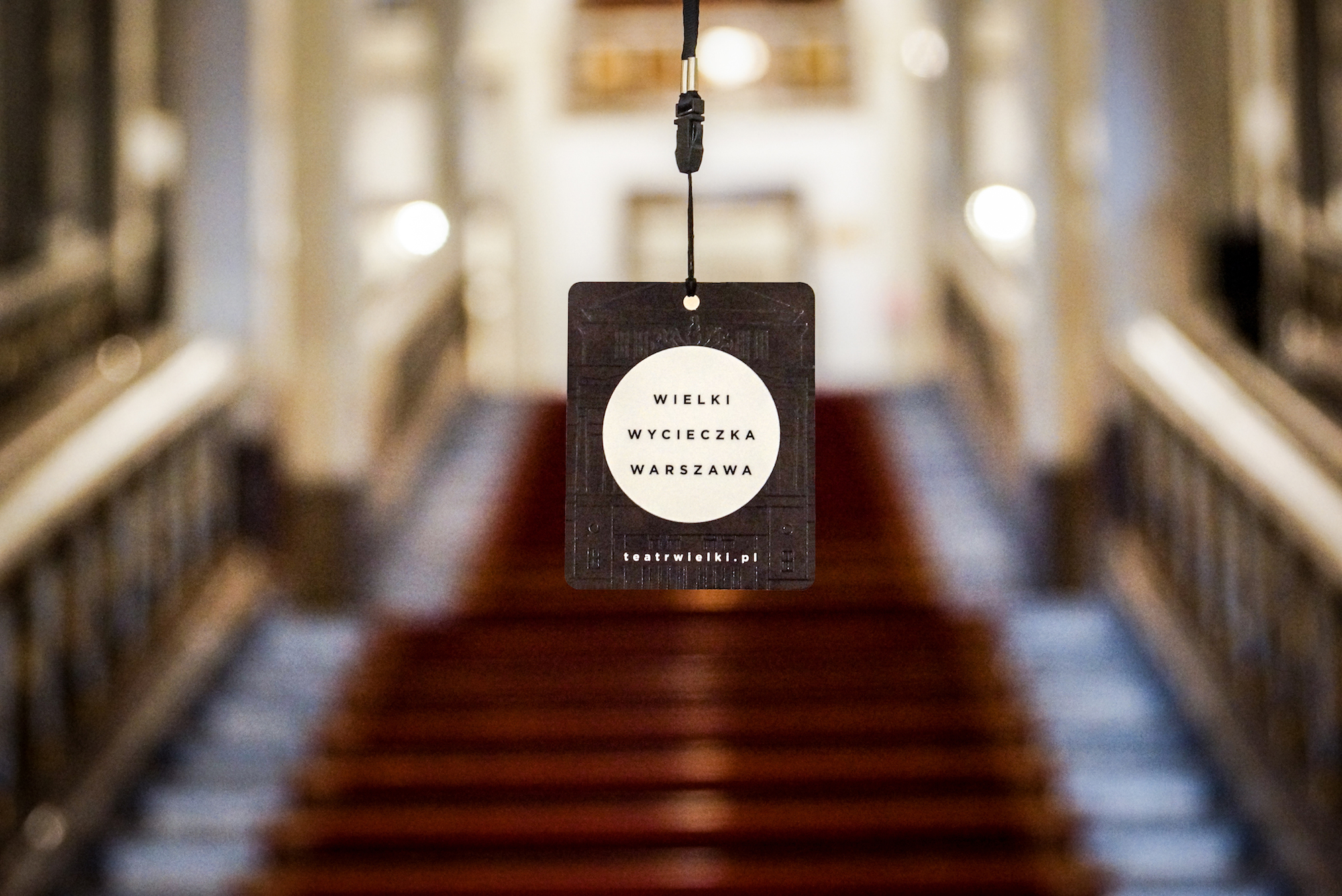 A badge reading 'Wielki, Tour, Warsaw’ with the red-carpeted main staircase at the Teatr Wielki