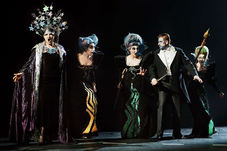 Four female singers in fantastic costumes, including elaborate headdresses, and a male singer in tails performing on the stage