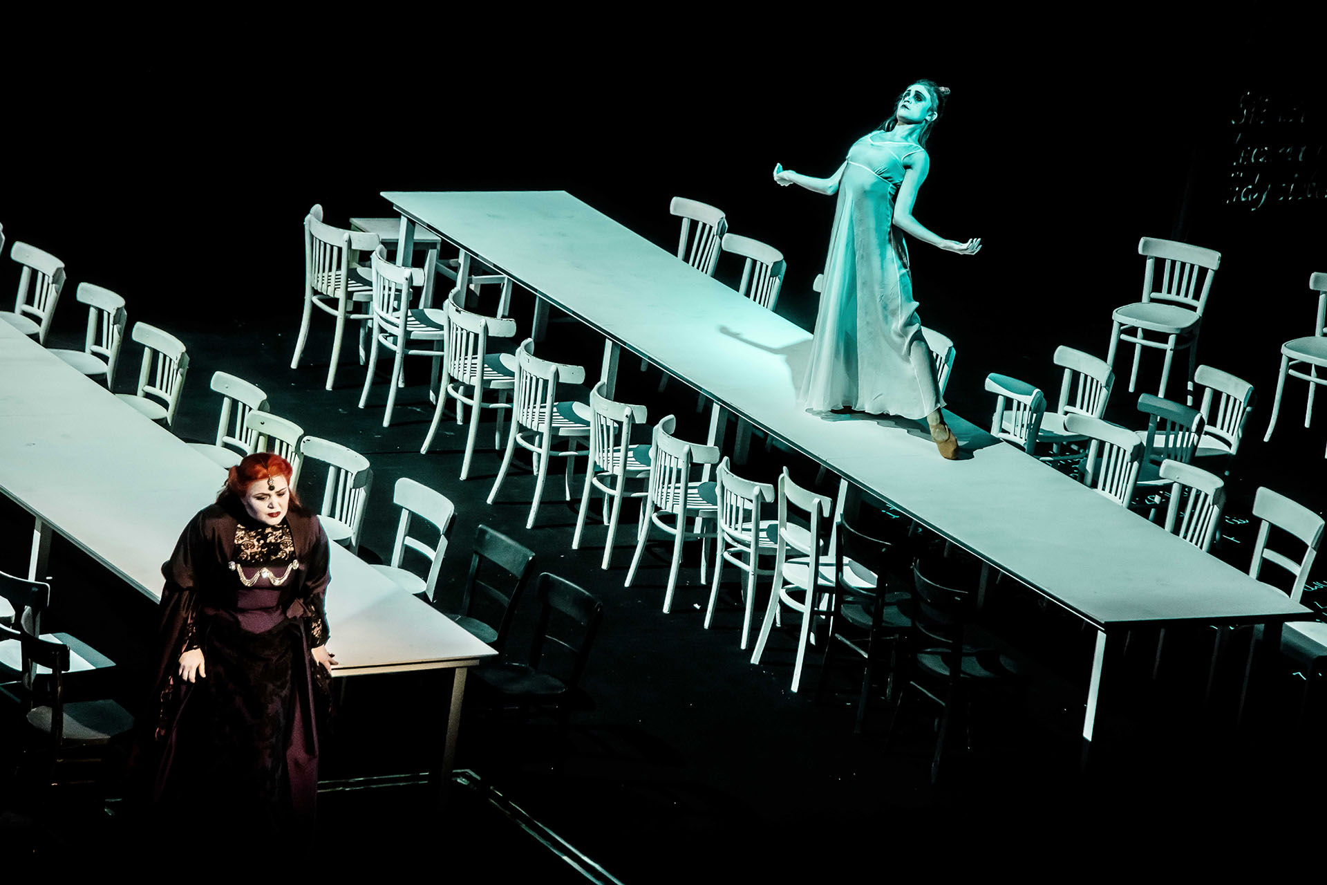 Na zdjęciu dwa długie stoły, przy nich krzesła. Na jednym ze stołów tancerka w zwiewnym kostiumie. Obok stoi śpiewaczka w roli Balladyny.