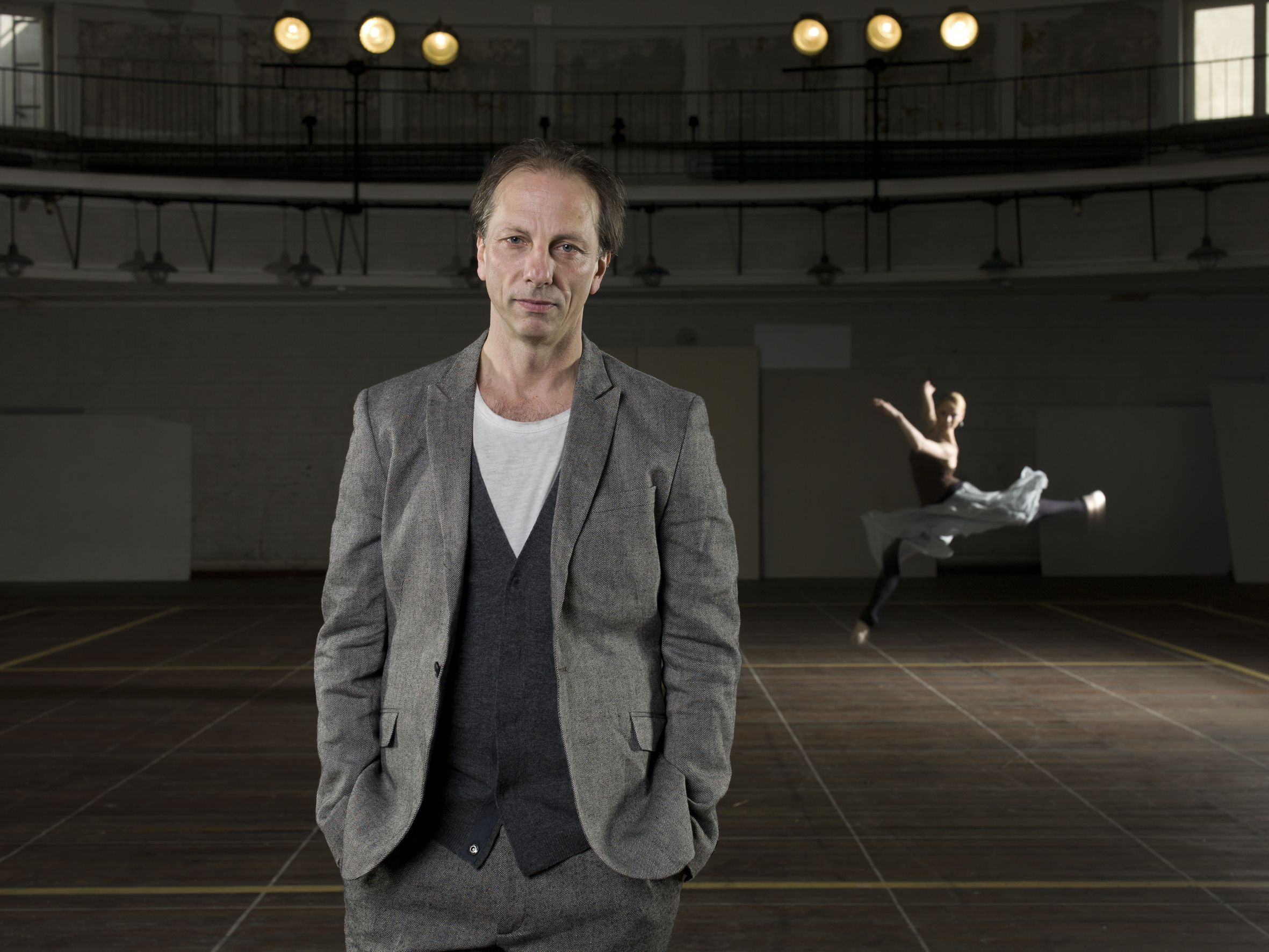 In the foreground, the artist in a gray suit, hands in his; behind his back, a female dancer mid-air in a jump
