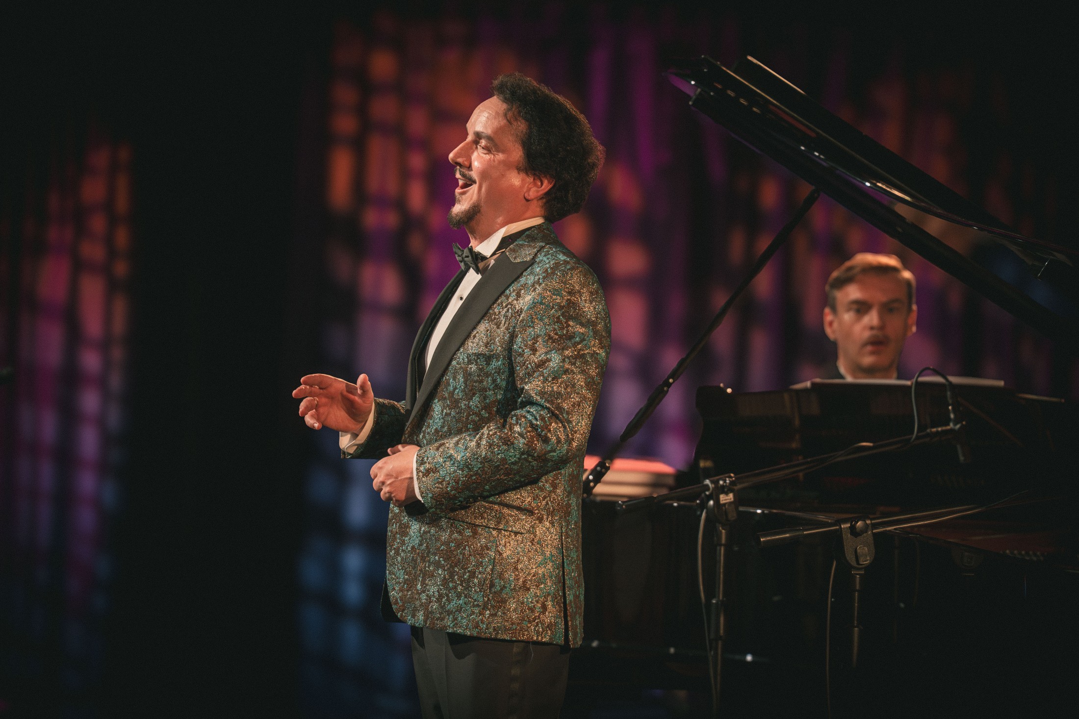 fotografia śpiewaka na scenie podczas koncertu, w tle pianista przy fortepianie