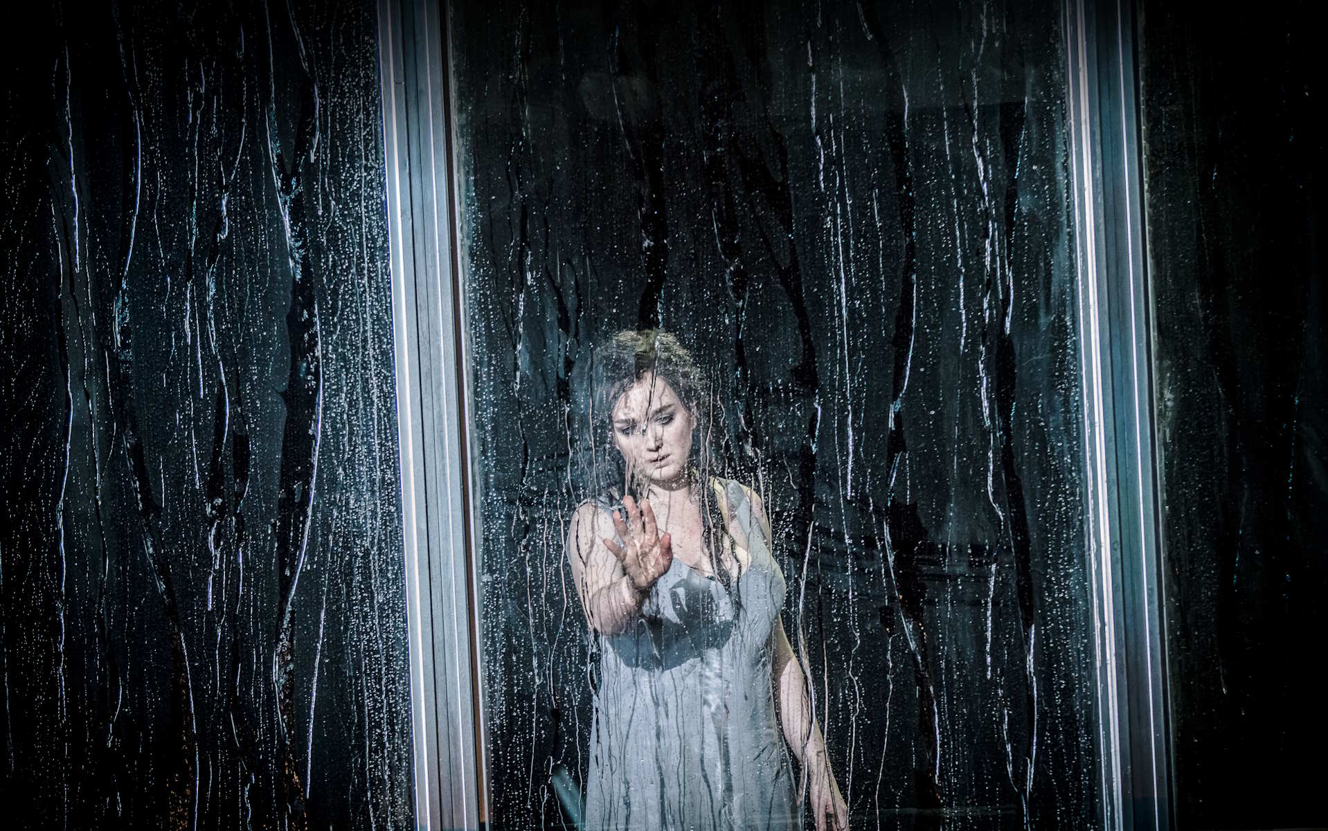 Kobieta ubrana w halkę dotyka ręką szyby, po której płyną strugi wody