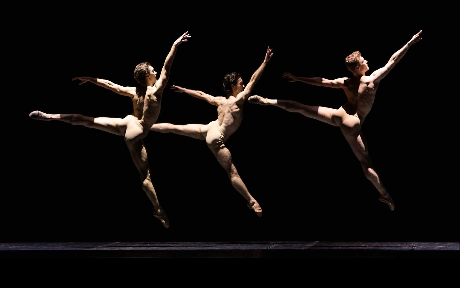 trzech tancerzy w cielistych kostiumach w jednakowym skoku, zwróceni w tę samą stronę
