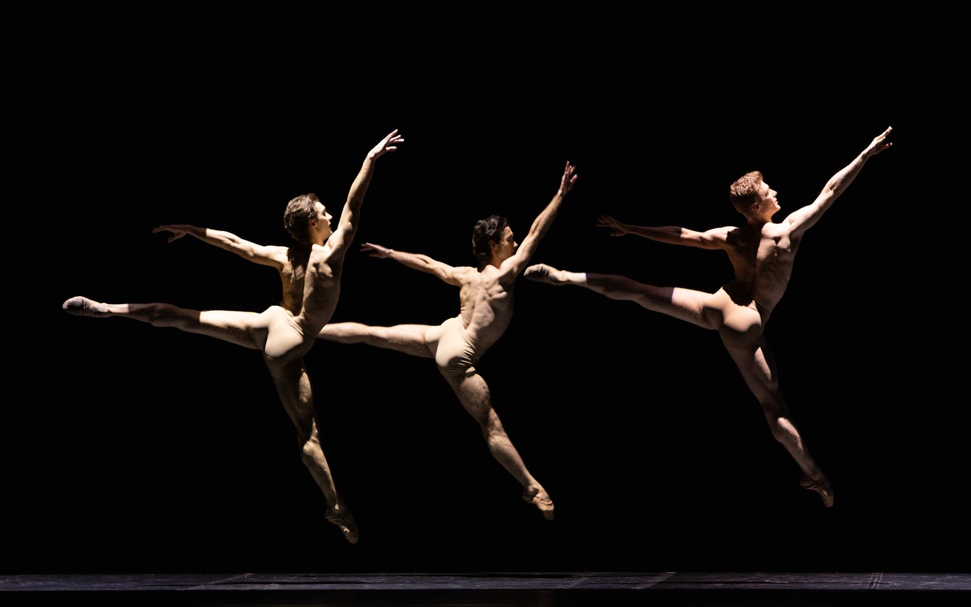 trzech tancerzy w cielistych kostiumach w jednakowym skoku, zwróceni w tę samą stronę